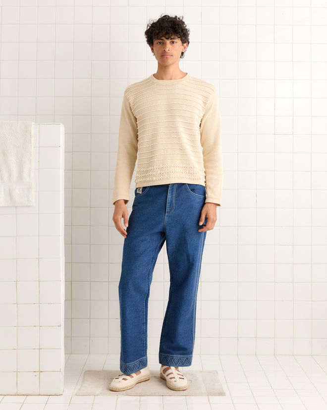 A person stands in a tiled room wearing a beige Beaded Pullover, blue jeans, and white shoes.