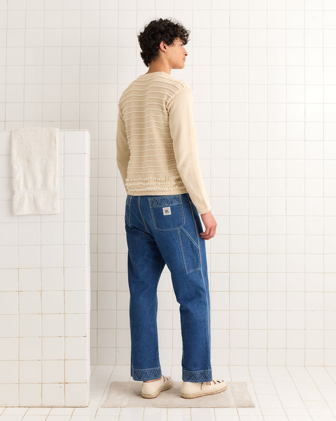 A man in blue jeans and the Beaded Pullover stands in a white-tiled bathroom, facing away.