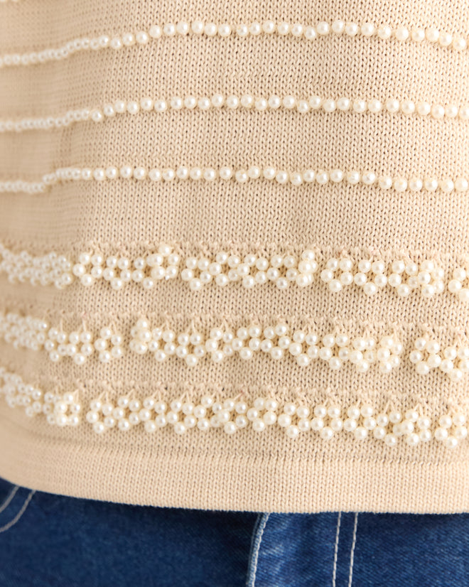 The Beaded Pullover, a cream hand-knit sweater embellished with rows of pearl-like beads, pairs effortlessly with blue denim jeans.