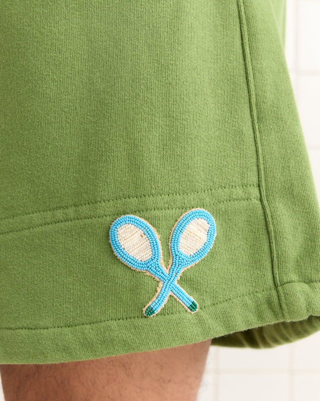 The Beaded Racquet Patch Shorts are green sweatshorts inspired by Canadian rugby style, featuring a beaded tennis patch with two crossed blue rackets embroidered near the hem.