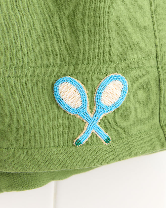 The Beaded Racquet Patch Shorts feature playful green fabric adorned with novelty tennis patches—two crossed rackets embroidered in blue and white beads—making them a fun addition to any outfit.
