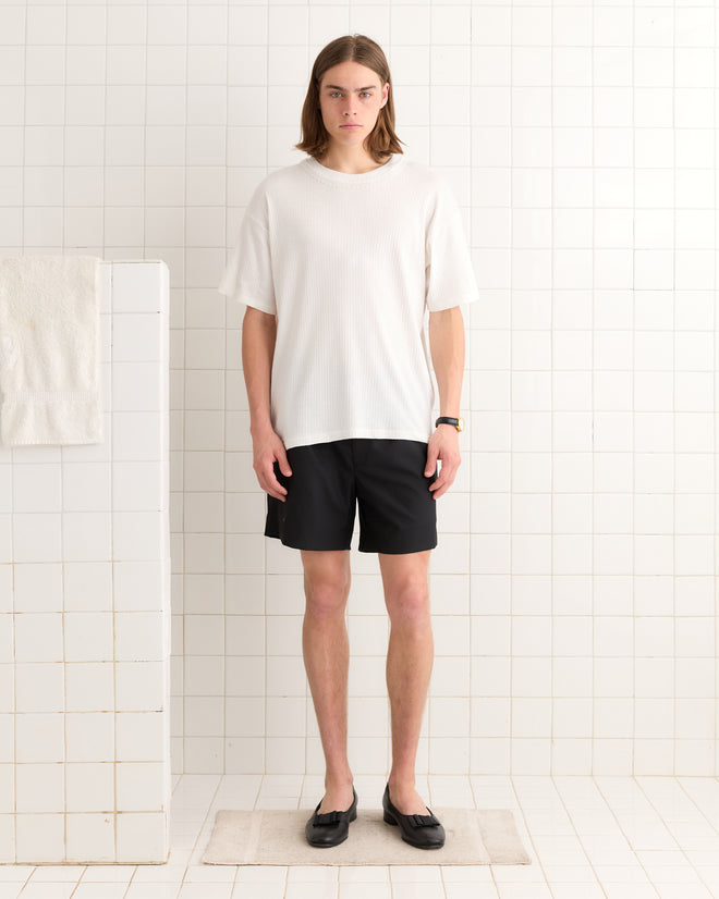 A person in a Beaded Salt Marsh Tee and black shorts stands on a white tiled floor, with a towel casually draped to the left.