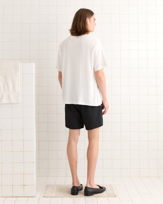 A person with medium-length hair in a Beaded Salt Marsh Tee and black shorts stands in a tiled room, facing away.