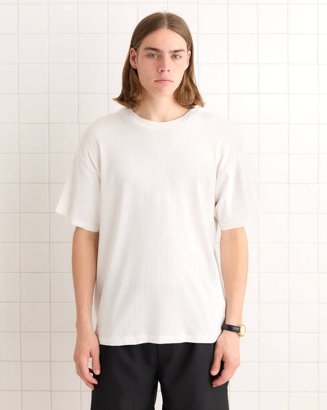 A person with long hair wearing a tonal white Beaded Salt Marsh Tee and black pants stands against a tiled wall.