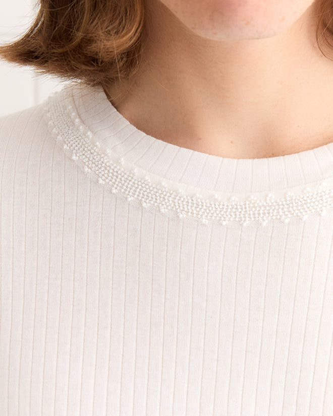 Close-up of a person wearing the Beaded Salt Marsh Tee, a tonal white ribbed cotton shirt featuring intricate beadwork around the neckline. Partial face visible.