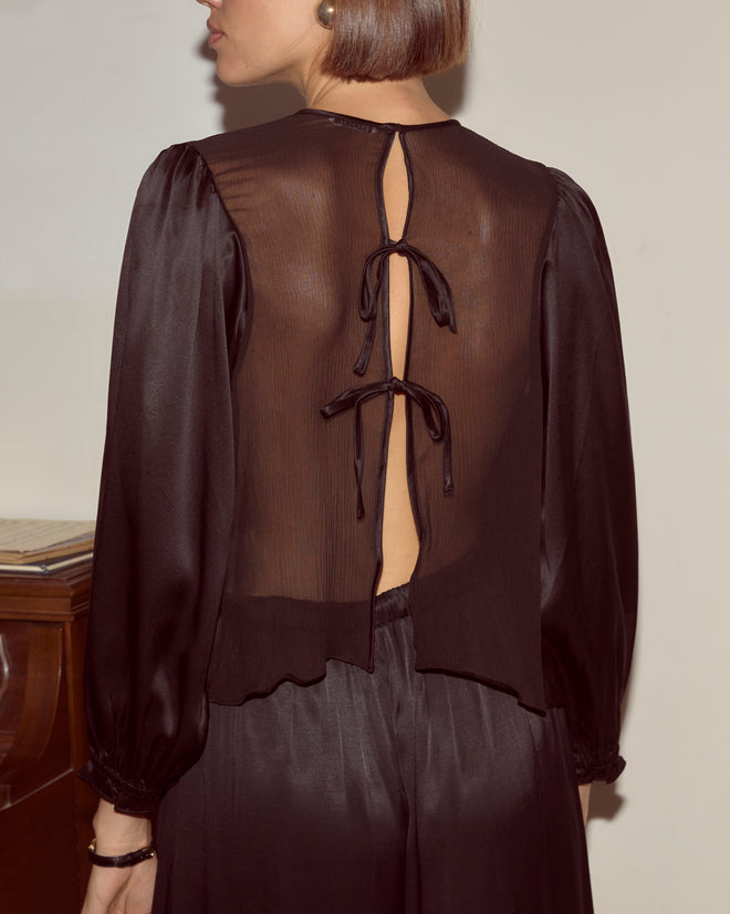A woman stands by a piano wearing the Belle Blouse - Black, a sheer silk blouse with an open back tied by two bows and delicate traditional shirring.