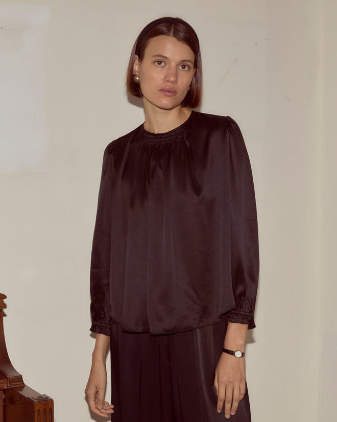 Woman with straight brown hair wears the Belle Blouse - Black, a loose silk top with traditional shirring, paired with a watch, standing against a plain beige wall.