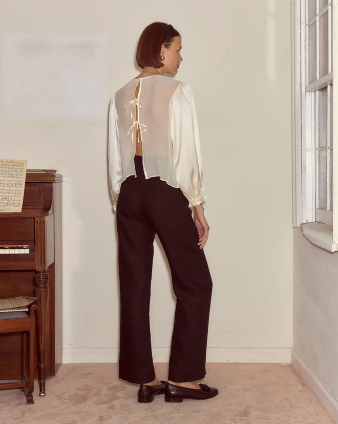 A woman stands indoors, facing away, wearing black pants and the Belle Blouse - Cream with traditional shirring tied at the back.