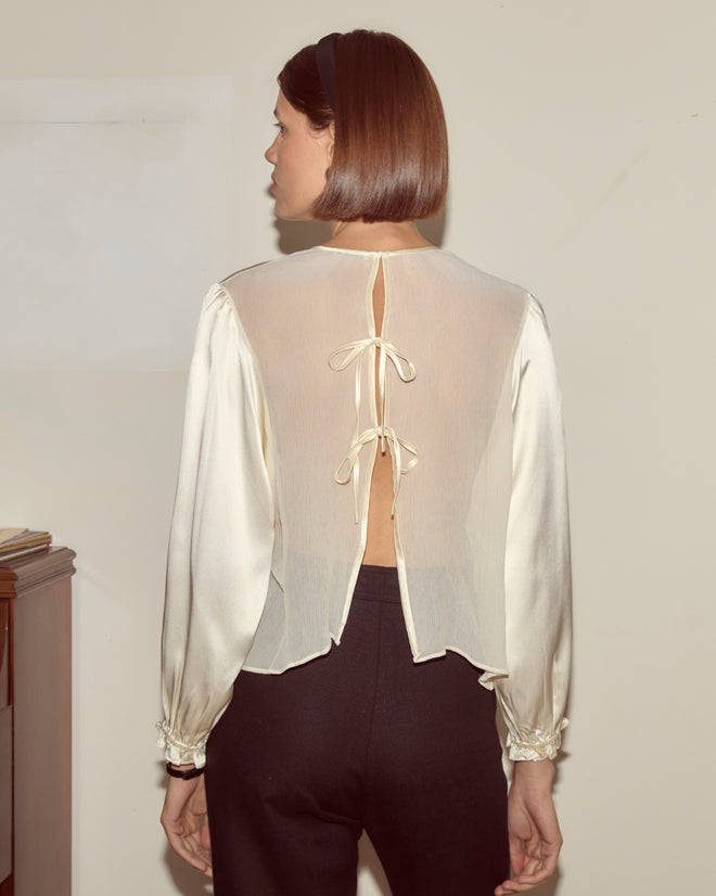 A woman stands indoors facing away, wearing the Belle Blouse in cream—a sheer silk folk-inspired top with traditional shirring and two back bows—paired with black pants.