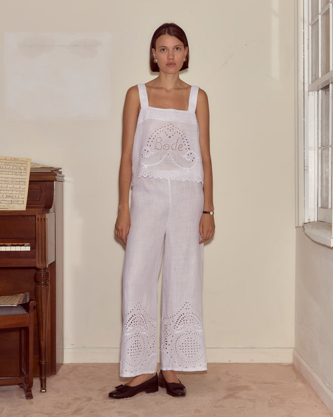 A woman stands indoors by a piano and window, looking at the camera while wearing Belvoir Pants and a white eyelet lace top.