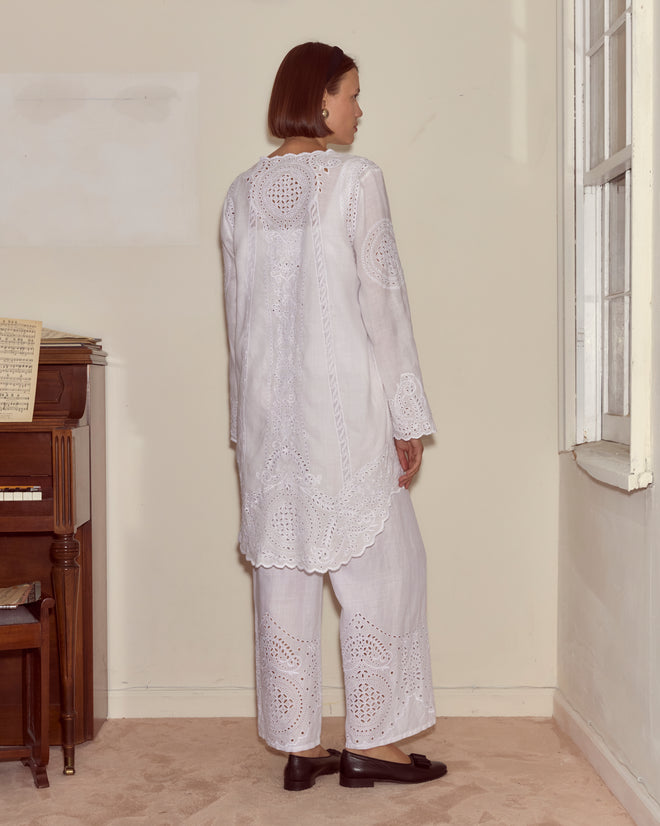 A woman stands near a piano, facing away, in a softly lit room with a window, wearing the Belvoir Top—a white broderie anglaise lace historical reproduction blouse.