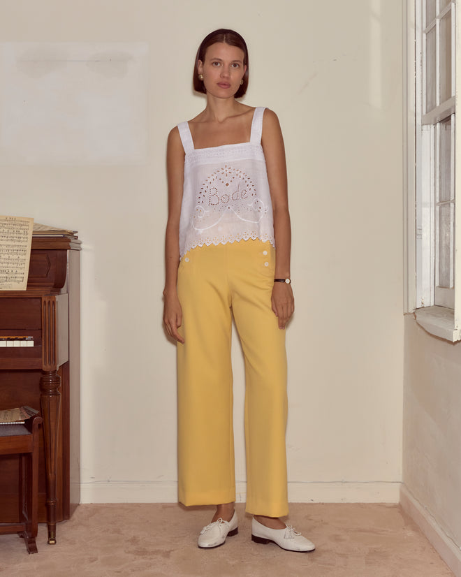A woman stands indoors by a piano and window, stylishly pairing yellow pants with the Belvoir Tank—crafted from 100% linen and finished with broderie anglaise lacework.