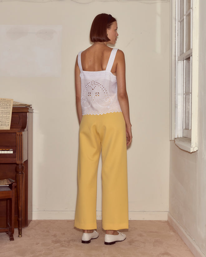 A woman with short hair stands facing a wall in a sunlit room, wearing the Belvoir Tank in white and yellow pants.