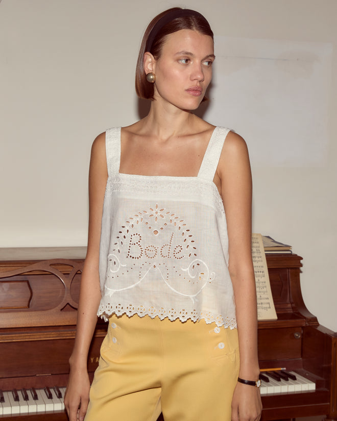 Woman wearing the Belvoir Tank, a vintage-inspired broderie anglaise linen top, paired with yellow pants stands in front of a piano.