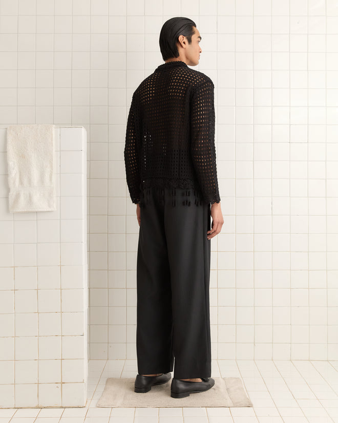 A person in a Blossom Crochet Shirt - Black and trousers stands elegantly in a tiled room adorned with a crocheted flower, with a towel hanging on the left wall.