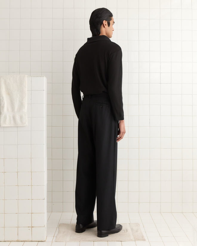 A person in the Boxy Long Sleeve Polo - Black stands on a tiled floor facing a white wall with a towel hanging. Their attire hints at 1970s fashion, possibly enhanced by the dry crepe yarn texture.