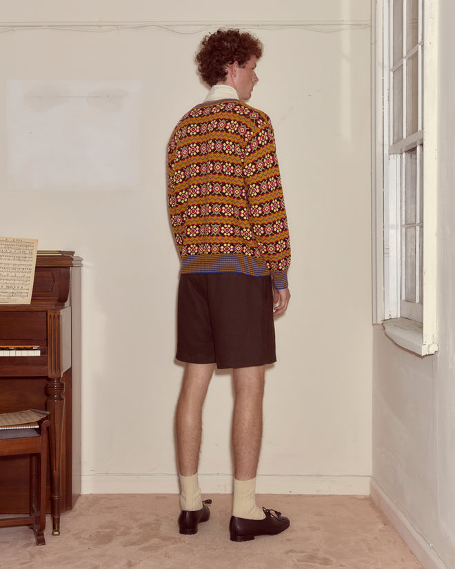 A man stands facing the corner in a vintage room, wearing the Bracken Sweater, dark shorts, socks, and loafers.