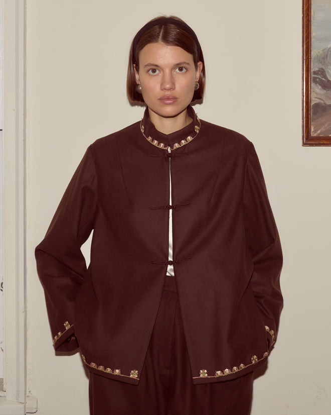 A woman stands indoors facing the camera with her hands in the pockets of a brown Bramble Jacket featuring gold trim.