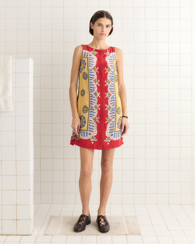A person wearing a Bricolage Dress, inspired by 1960s patterns in red, stands on a mat in a tiled room with black shoes.