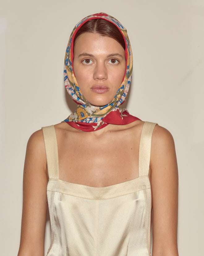 A woman in a cream dress and colorful headscarf stands before a beige backdrop, channeling vintage charm with the Bricolage Driving Cap.