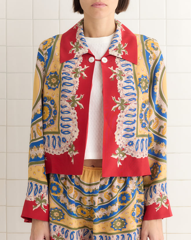 A person in a vibrant, paisley-patterned outfit featuring red, yellow, and blue stands against a tiled wall. Their look is elevated by the exquisite Bricolage Jacket adorned with antique pearl buttons.