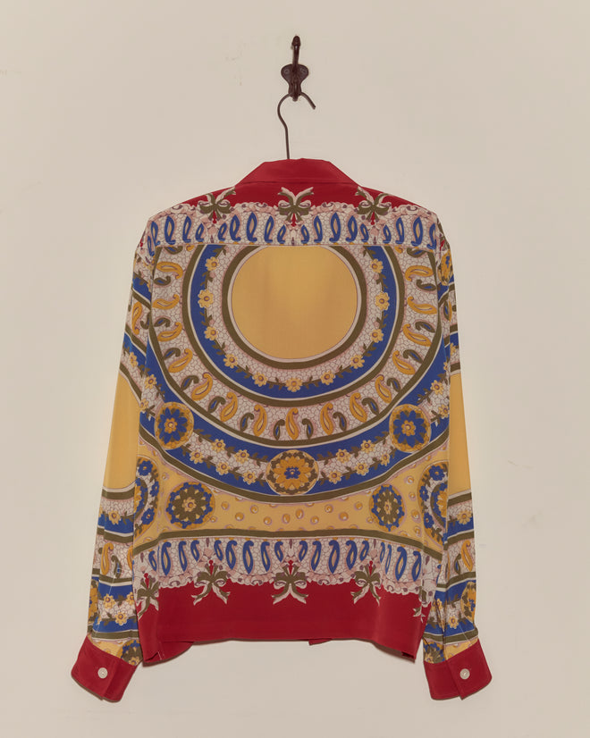 The Bricolage Long Sleeve Shirt, a 1960s-inspired silk top with red, yellow, blue, and gold patterns, hangs on a hook against a white wall.