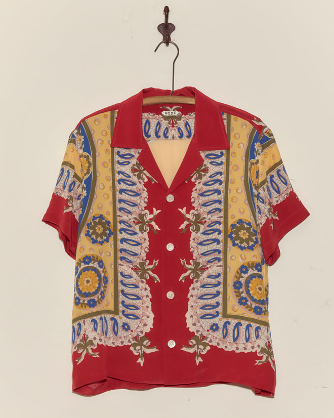 The Bricolage Short Sleeve Shirt, a 1960s-inspired red silk top with vibrant yellow, blue, and floral patterns, hangs on a hook against a white wall.
