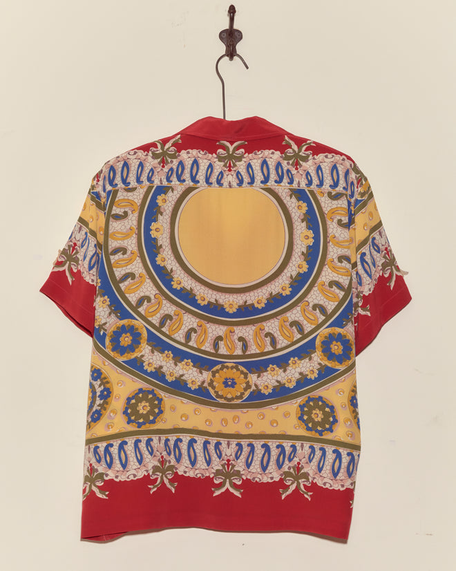 The Bricolage Short Sleeve Shirt, featuring colorful yellow, blue, and red patterns on silk, hangs on a hook against a wall.