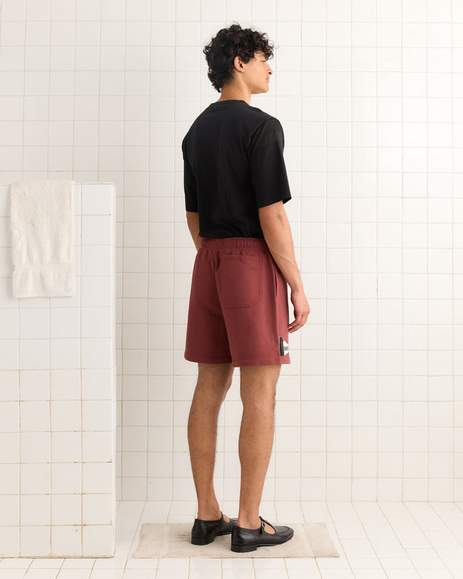 A person stands on a mat in a tiled room, wearing a black shirt, Burgundy Pennant Shorts, and black shoes.