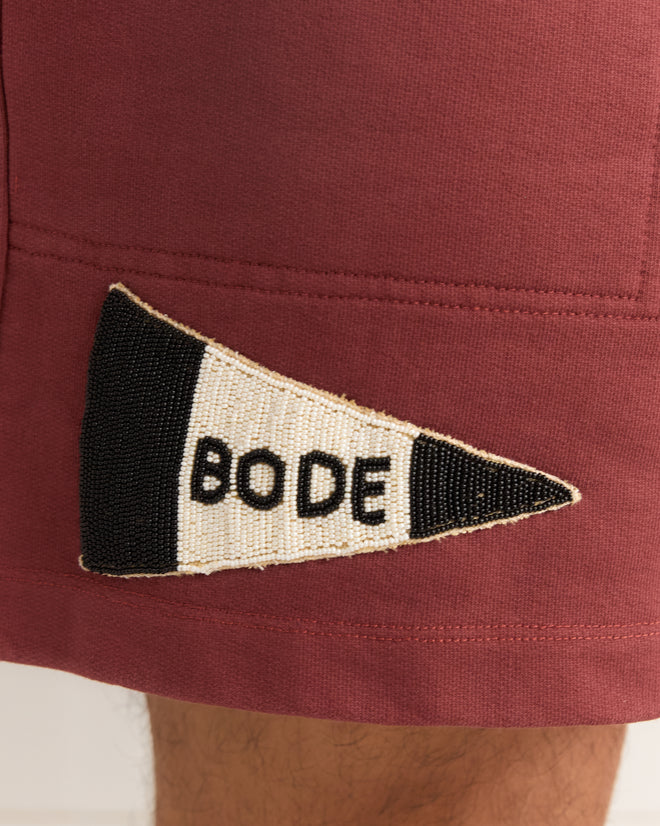 The Burgundy Pennant Shorts feature maroon cotton fabric and a beaded “BODE” patch in black letters on a white and black triangle, inspired by classic rugby shorts.