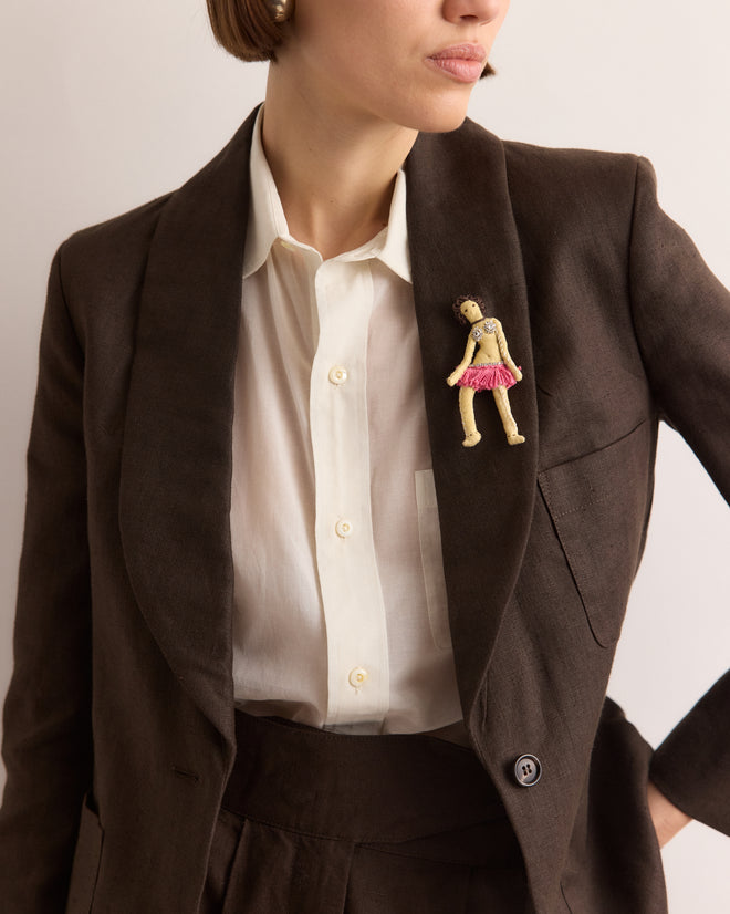 Woman in a brown suit and white shirt wears the Calder Fanni Brooch, inspired by Alexander Calder’s jewelry and shaped like a doll in a pink skirt, reminiscent of the Calder Circus brooch from the Whitney Museum collection.