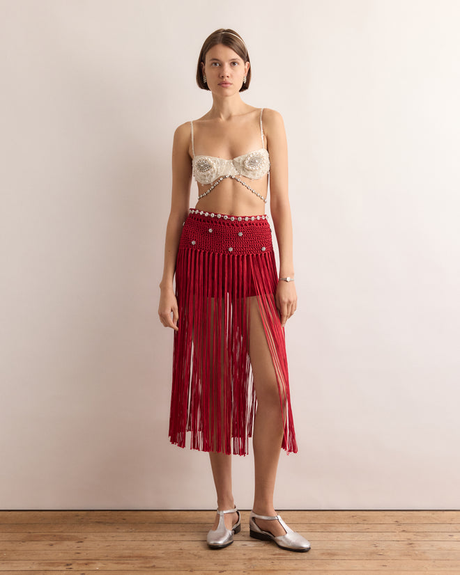 Woman in the Calder Fanni Fringe Skirt, featuring red fringe and a thigh slit, stands on a wooden floor against a plain wall—channeling the playful spirit of Calder’s Circus as seen at the Whitney Museum of American Art.