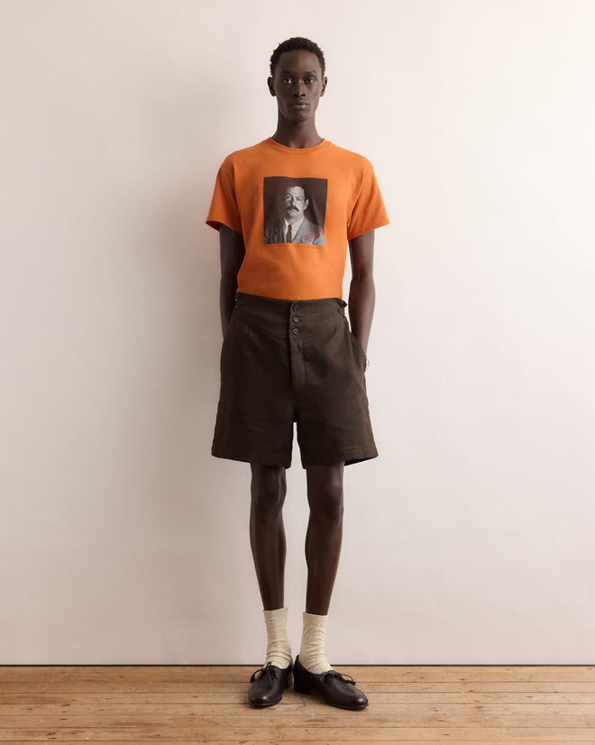 A person wears the Calder Identity Card Tee—an orange graphic T-shirt—paired with brown shorts, cream socks, and black loafers against a plain background.