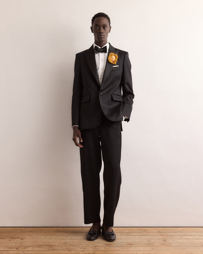 A person in a black tuxedo with a yellow flower on the lapel stands against a plain white background, elegantly accessorized with the Calder Leo Brooch.
