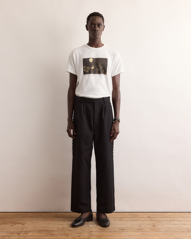 A person stands against a wall wearing the Calder Suitcase Tee, a white graphic T-shirt inspired by Alexander Calder’s Circus, paired with black pants and black shoes.