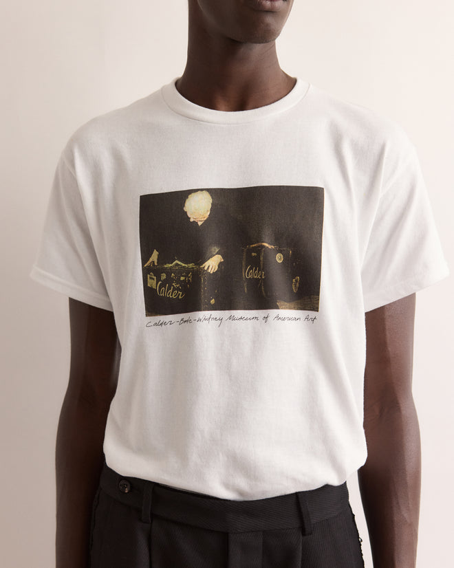 A person wearing the Calder Suitcase Tee, a white T-shirt with an abstract black and yellow graphic and cursive text inspired by Alexander Calder’s Circus, evoking the Whitney Museum of American Art.