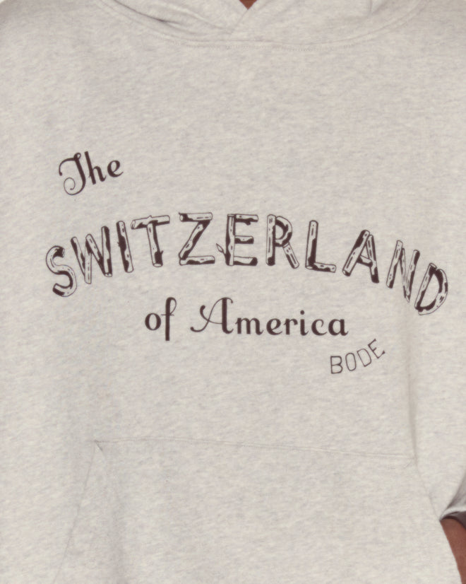 The Campground Hoodie is a light gray sweatshirt with “The SWITZERLAND of America BODE” and a subtle log cabin illustration on the front, adding signature Camp Geneva charm.