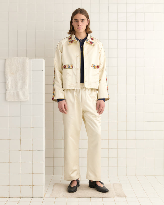 In a tiled room, someone in a Candyfloss Jacket with embroidered floral trim stands, while a towel hangs on a partition.