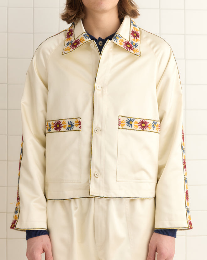 A person in a Candyfloss Jacket, featuring embroidered floral trim on the collar, pockets, and sleeves, stands gracefully before a tiled wall.