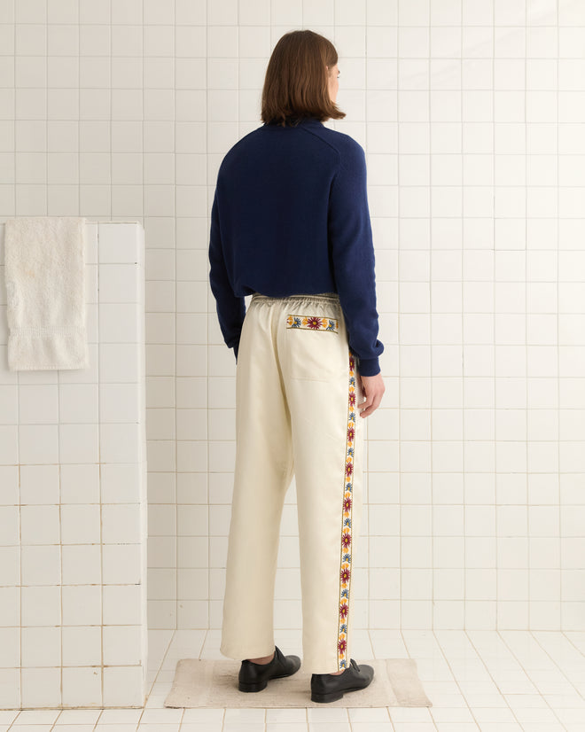 In a tiled room, a person in Candyfloss Track Pants with colorful yarn embroidery on the side stripes, navy sweater, and cream pants stands facing away.