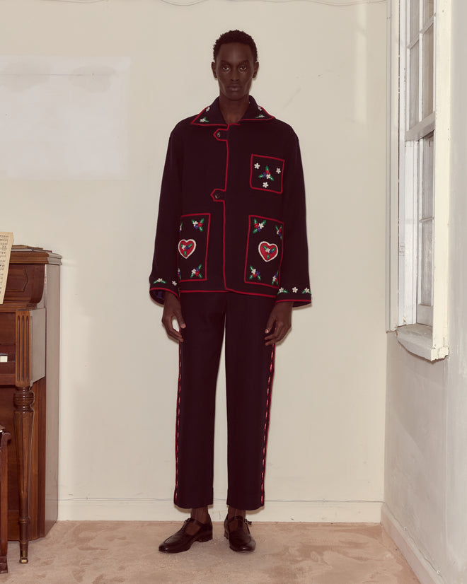 A person stands indoors near a piano and window, wearing the Carroll Front Tab Jacket—a dark embroidered wool piece with heart and floral details, evoking an Alice Through the Looking Glass aesthetic.