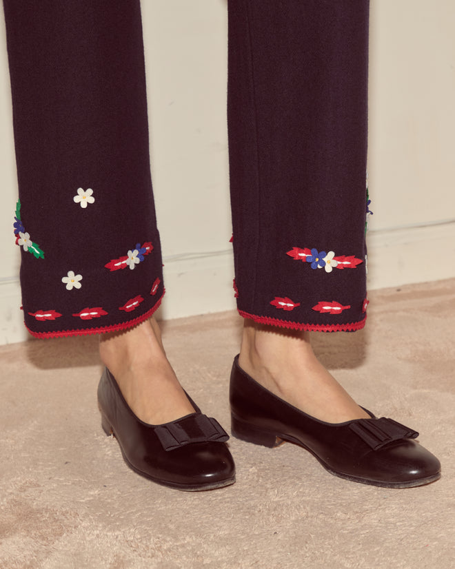 Wearing Carroll Trousers with floral embroidery near the hem and black bow flats, the person stands on a beige carpet.