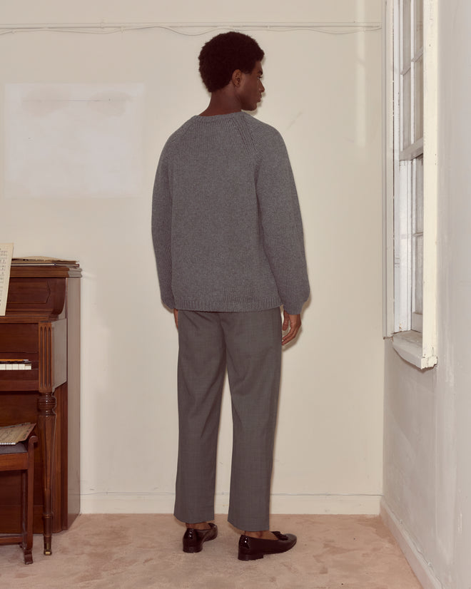Wearing the Cashmere Crewneck - Grey with an oversized fit, a person stands facing a corner in a room featuring a piano and a window.
