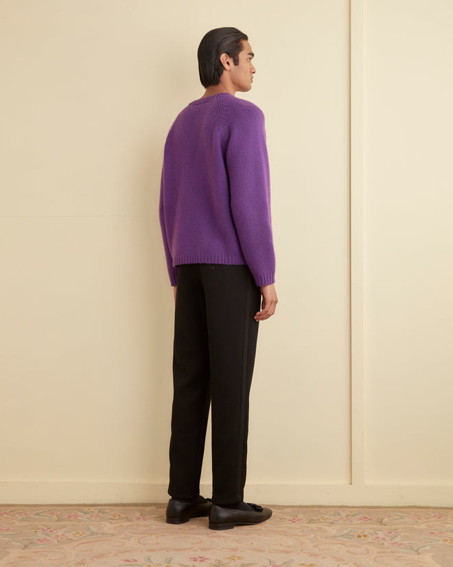 A person in an oversized purple Cashmere Crewneck Sweater and black pants stands against a beige wall on a patterned carpet.