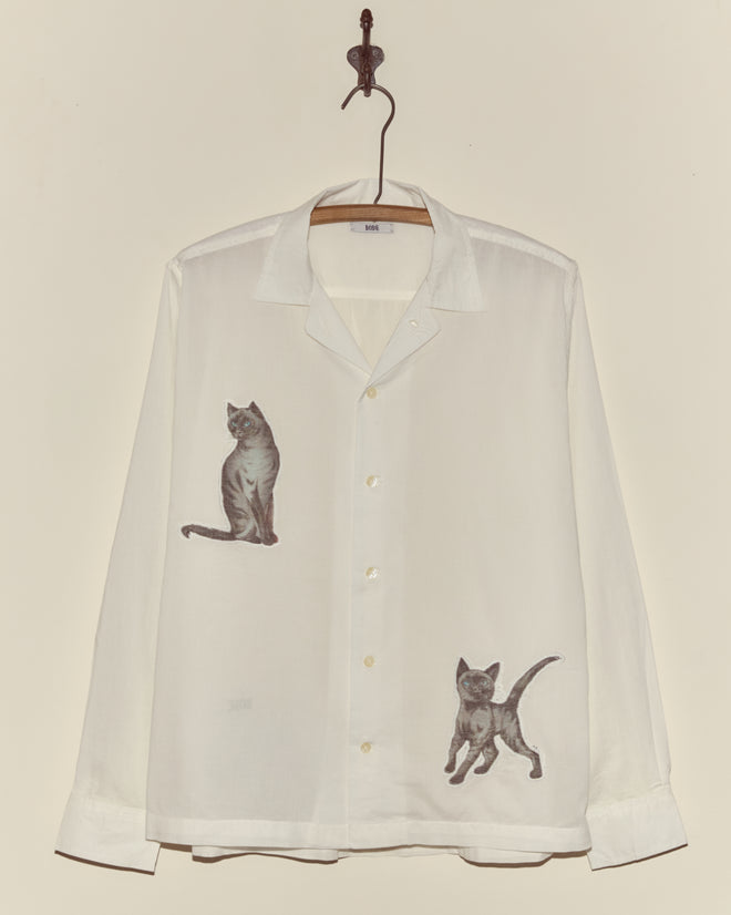 The Cat Motif Long Sleeve Shirt is a white, vintage-inspired button-up featuring illustrations of two playful gray cats, displayed on a hook against a plain wall.