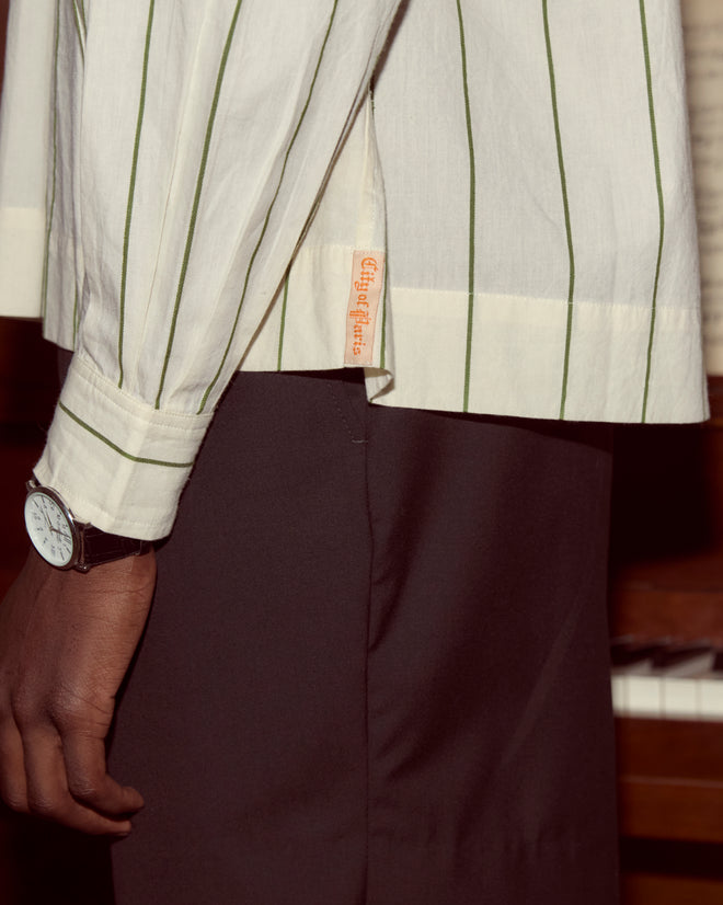 A person wearing the Celery Stripe Cricket Shirt, black pants, and a watch stands near a piano keyboard.