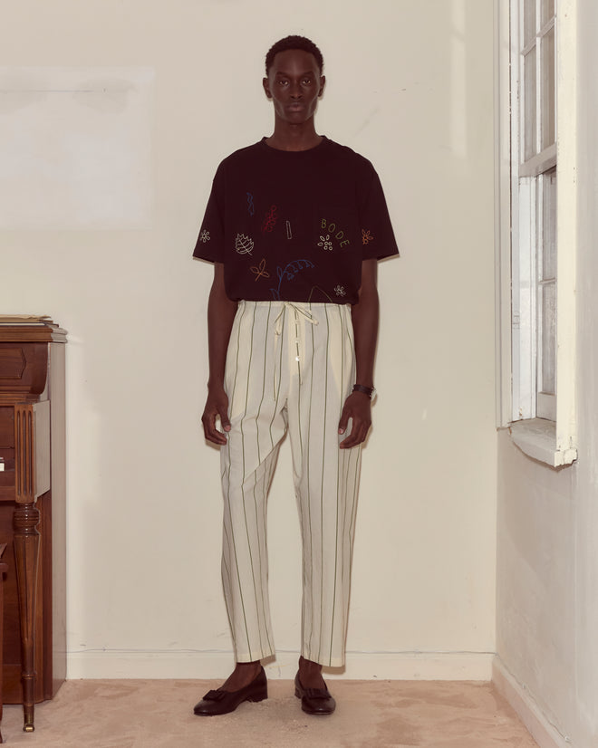A person stands indoors near a window and piano, wearing a black t-shirt with designs and Celery Stripe Pajama Pants.