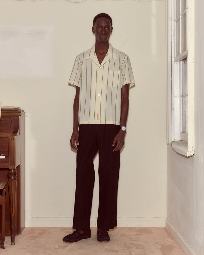A man stands indoors wearing the Celery Stripe Short Sleeve shirt with an antique City of Paris label, paired with black pants and brown shoes.