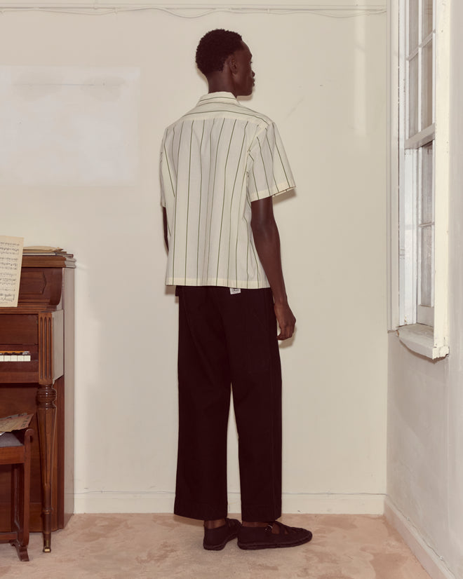 A person stands indoors near a piano and window, wearing the Celery Stripe Short Sleeve vintage shirt made of 100% cotton with black pants, facing a wall.