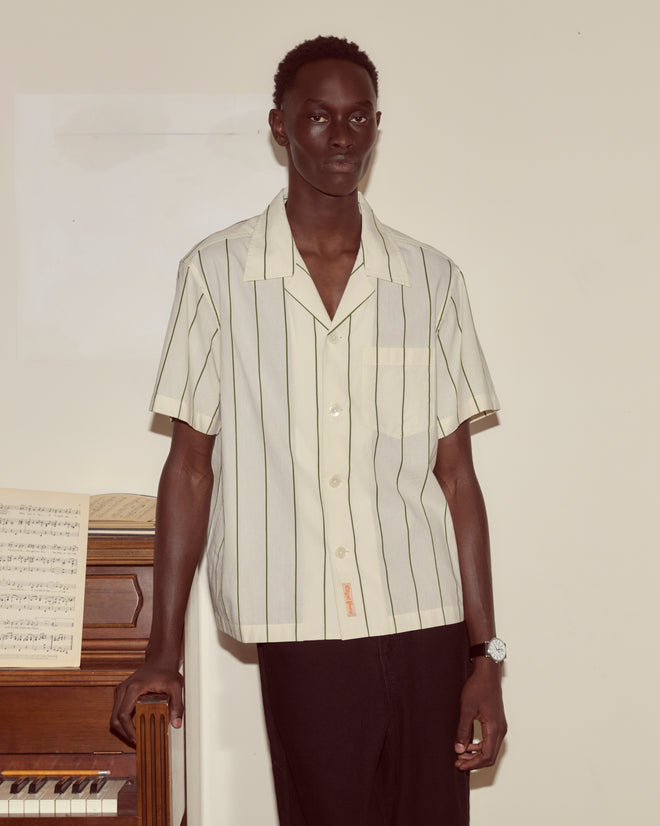 A man in a Celery Stripe Short Sleeve shirt with an antique City of Paris label stands by a piano with sheet music, looking at the camera.